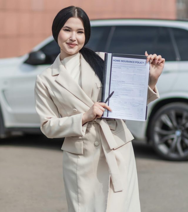 Smiling woman in beige blazer holding a home insurance policy document in outdoor setting.